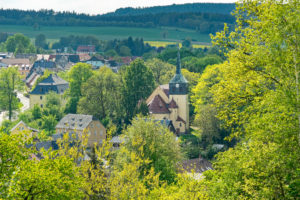 Neusalza-Spremberg in der Oberlausitz mit bunten Häusern und dem hohen Kirchturm der Spremberger Kirche, umgeben von üppig grünen Bäumen und sanften Hügeln unter einem teilweise bewölkten Himmel.