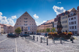 Ein sonniger Stadtplatz mit Kopfsteinpflaster, parkenden Autos, bunten historischen Gebäuden und dem berühmten Salzhaus in Zittau. Ein Gebäude mit dreieckigem Dach steht in der Nähe eines dekorativen, mit Blumen gefüllten Springbrunnens, während die Menschen in der Nähe spazieren gehen und sich versammeln.