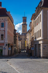 Eine Kopfsteinpflasterstraße in Zittau, gesäumt von historischen Gebäuden, führt zu einem hohen, verzierten Uhrenturm unter einem strahlend blauen Himmel mit ein paar weißen Wolken.