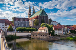 Eine malerische Ansicht der historischen Stadt Görlitz mit farbenfrohen Gebäuden, der großen Peterskirche mit Zwillingstürmen und einem grünen Dach, gelegen an der Neiße unter einem teilweise bewölkten Himmel. Menschen speisen auf einer Terrasse am Flussufer.