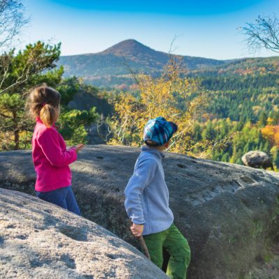 Zwei Kinder, eines in einer rosafarbenen Jacke und das andere in einer grauen Jacke mit blauem Hut, stehen auf großen Felsen im Zittauer Gebirge und blicken auf eine malerische Waldlandschaft mit der Lausche in der Ferne unter einem klaren blauen Himmel. Toll für Wanderungen mit Kindern.