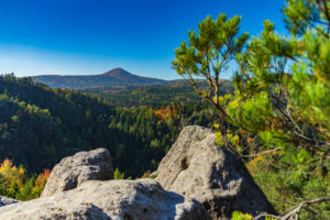 Felsvorsprung mit Blick auf ein bewaldetes Tal mit dichten grünen Bäumen und buntem Herbstlaub im Zittauer Gebirge. In der Ferne erhebt sich der Berg Lausche unter einem strahlend blauen Himmel, während ein belaubter Ast die rechte Seite einrahmt - perfekt zum Wandern.