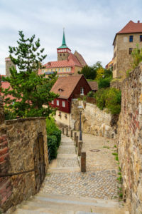 Ein kopfsteingepflasterter Weg, gesäumt von Steinmauern und Grünflächen, führt hinunter zu rot gedeckten Gebäuden und einer Kirche mit einer hohen, spitzen Turmspitze in der historischen europäischen Stadt Bautzen.