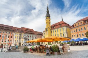 Die Menschen sitzen an Tischen im Freien unter großen gelben Sonnenschirmen auf einem farbenfrohen europäischen Stadtplatz. Im Hintergrund erheben sich pastellfarbene Gebäude und der hohe Uhrenturm des historischen Rathauses Bautzen unter einem teilweise bewölkten Himmel.