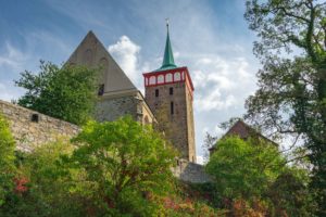 Eine historische Steinkirche in Bautzen mit rot-weißem Turm und grüner Spitze erhebt sich über einer Steinmauer, umgeben von üppig grünen Bäumen und Büschen unter einem teilweise bewölkten Himmel.