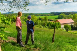 Zwei Personen stehen auf einem grasbewachsenen Hügel mit Blick auf grüne Felder, Bäume und ein großes Gebäude mit rotem Dach unter einem teilweise bewölkten Himmel - perfekt für einen Aktivurlaub voller Entspannung und Outdoor-Abenteuer in dieser friedlichen ländlichen Umgebung. Blick von den Schmiedesteinen aus in Richtung Neusalza-Spremberg.
