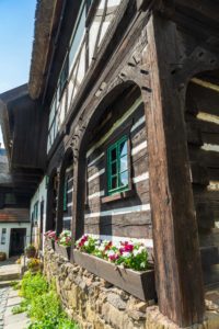 Das rustikale Reiterhaus in Neusalza-Spremberg mit Holzbalken, Steinfundament und Blumenkästen unter grün umrandeten Fenstern. Helle Blüten sonnen sich im Sonnenlicht und fangen den gemütlichen Charme der Oberlausitz ein.