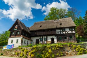 Das traditionelle Reiterhaus mit Reetdach steht auf einer Steinmauer in Neusalza-Spremberg, umgeben von üppigem Grün unter einem blauen Himmel mit vereinzelten Wolken.