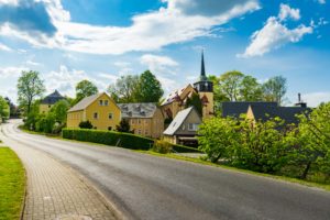 Eine ruhige, ländliche Kleinstadt mit gelben Häusern, einer Kirche mit einem hohen Kirchturm, Bäumen und einer kurvenreichen Straße unter einem teilweise bewölkten blauen Himmel bildet ein reizvolles Panorama von Neusalza-Spremberg.