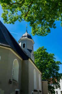 Die Neusalzaer Kirche mit ihren gewölbten Fenstern und dem mit einem Kreuz gekrönten Uhrenturm steht unter grünen Bäumen vor einem strahlend blauen Himmel.