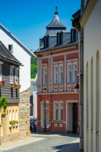 Eine schmale, kopfsteingepflasterte Straße in Neusalza-Spremberg mit historischen Gebäuden, darunter ein rosafarbenes und weißes Eckhaus mit verzierten Details und einem Turmdach, unter einem strahlend blauen Himmel.