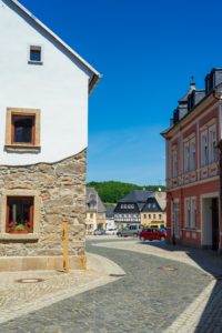 Eine kopfsteingepflasterte Straße schlängelt sich zwischen historischen Stein- und Stuckgebäuden mit bunten Fassaden. Ein paar Autos sind entlang der Straße geparkt, und im Hintergrund sind grüne Bäume und blauer Himmel zu sehen.