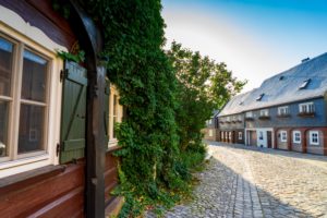 An einem sonnigen Tag schlängelt sich eine Kopfsteinpflasterstraße zwischen traditionellen Umgebindehäusern in der Oberlausitz, von denen eines mit grünen Weinreben bewachsen ist. Die Szenerie ist friedlich, der Himmel klar und blau und die Blumenkästen an den entfernten Gebäuden.