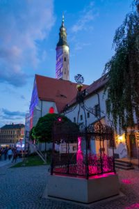 Die Klosterkirche Zittau mit ihrem hohen Kirchturm steht hinter einem kleinen, kunstvoll verzierten, schmiedeeisernen Pavillon, der in der Abenddämmerung mit rosa Lichtern beleuchtet wird, während die Menschen eine von historischen Gebäuden gesäumte Kopfsteinpflasterstraße entlangschlendern.
