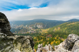 Blick vom Scharfenstein auf den Berg Oybin. Ein üppiges, grünes Tal mit dichtem Wald, vereinzelten Herbstfarben und einem entfernten Dorf inmitten von Bergen unter einem teilweise bewölkten Himmel.