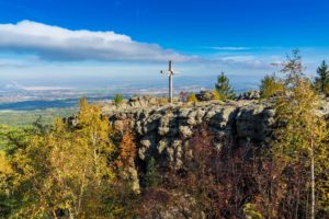Ein großes Holzkreuz steht auf einem Felsen im Zittauer Gebirge, umgeben von herbstlichen Bäumen, mit Blick auf ein fernes Tal und eine Stadt unter einem strahlend blauen Himmel mit vereinzelten Wolken - ein perfekter Ort zum Wandern.