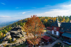 Ein malerischer Blick auf das Zittauer Gebirge im Herbst, mit bunten Bäumen, einem Felsvorsprung und dem roten Dach der Töpferbaude unter blauem Himmel. Menschen gehen spazieren und genießen die malerische Landschaft im Freien.