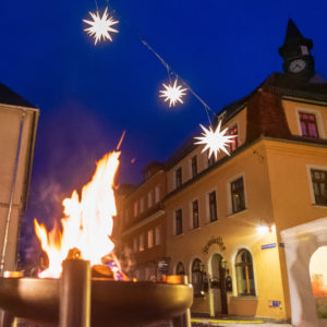 Während des Weihnachtsmarktes in Neusalza-Spremberg brennt ein Feuer in einem metallenen Kohlenbecken auf dem Obermarkt. Im Hintergrund ist ein gelbes Gebäude mit einem Uhrenturm zu sehen, darüber befinden sich dekorative Herrnhuter Sterne.