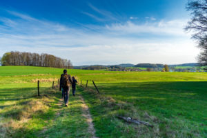 Zwei Personen mit Rucksäcken wandern auf dem Oberlausitzer Bergweg, einem unbefestigten Weg durch eine grüne Wiese unter einem blauen, leicht bewölktem Himmel, der sich perfekt zum Wandern oder für einen Familienausflug inmitten von fernen Bäumen und sanften Hügeln eignet.