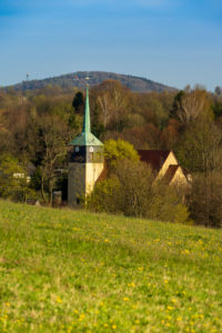 Eine ländliche Szene mit einer Kirche, deren grüner Turm von Bäumen umgeben ist, einem abfallenden Grasfeld mit gelben Blumen im Vordergrund und bewaldeten Hügeln unter blauem Himmel im Hintergrund.