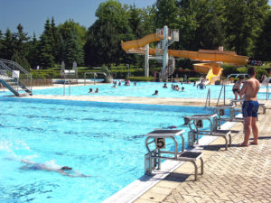 Schwimmen und Entspannen im Wald- und Erlebnisbades Neusalza-Spremberg ein Freibad mit Schwimmbahnen, Sprungtürmen und einer von Bäumen umgebenen gelben Wasserrutsche. Ein Mann in Badehose steht in der Nähe des Beckenrandes.