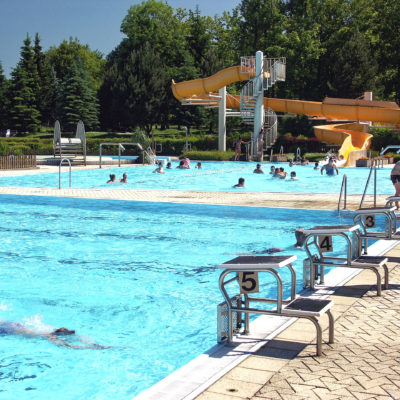 Schwimmen und Entspannen im Wald- und Erlebnisbades Neusalza-Spremberg ein Freibad mit Schwimmbahnen, Sprungtürmen und einer von Bäumen umgebenen gelben Wasserrutsche. Ein Mann in Badehose steht in der Nähe des Beckenrandes.