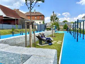 Eine Person sitzt auf einer geschwungenen Bank in einem modernen Sportpark in Neusalza-Spremberg mit blauen Wegen, grünem Gras, jungen Bäumen und nahegelegenen Wohngebäuden unter einem teilweise bewölkten Himmel und entspannt sich.