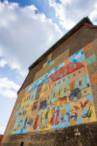Ein Wandgemälde an der Fassade eines Gebäudes in Neusalza-Spremberg zeigt eine lebendige Straßenszene mit bunten Häusern, Menschen, Pferden und verschiedenen Tieren, alles unter einem hellen, teilweise bewölkten Himmel.