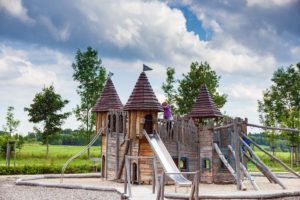 Ein hölzernes Spielgerät in Form einer Burg mit Rutschen und Türmen steht an einem bewölkten Tag im Freien. Auf dem Spielplatz Friedersdorf in der Oberlausitz spielt ein Kind ganz oben, umgeben von Bäumen und grünen Wiesen - ein perfektes Wanderziel für Familien.