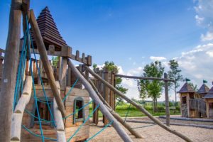 Der Spielplatz in Friedersdorf besteht aus einer hölzernen Spielplatzstruktur mit Kletterseilen und Turmdächern, die auf einer Kiesfläche steht. Im Hintergrund sind grünes Gras, Bäume und ein teilweise bewölkter blauer Himmel zu sehen.