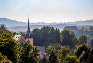 Eine Kirche mit einem hohen Kirchturm erhebt sich über den Bäumen in der üppigen, grünen Landschaft des Oberlausitzer Berglandes bei Neusalza-Spremberg, mit sanften Hügeln und einem nebligen Himmel im Hintergrund.
