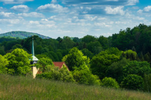 Eine kleine Kirche in der Oberlausitz mit grünem Kirchturm und rotem Dach liegt inmitten üppiger grüner Bäume auf einem Hügel, mit einem grasbewachsenen Feld im Vordergrund und entfernten Hügeln unter einem teilweise bewölkten Himmel.