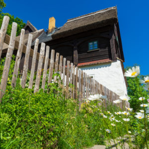 Hinter einem Lattenzaun steht ein rustikales Holzhaus mit Strohdach, das Reiterhaus Neusalza-Spremberg. Im Vordergrund wachsen Wildblumen und hohes Gras unter einem strahlend blauen Himmel.