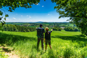 Zwei Wanderer stehen auf einer grasbewachsenen Hügelkuppe, umgeben von üppigem Grün, und blicken auf die malerische Landschaft in der Nähe von Ferienwohnungen in der Oberlausitz in Sachsen mit Feldern, Bäumen und fernen Hügeln unter einem klaren blauen Himmel. Ein Wanderer zeigt auf die atemberaubende Aussicht.