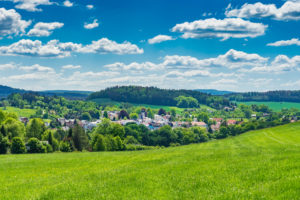 Eine malerische Ansicht von Neusalza-Spremberg, einer kleinen Stadt inmitten der grünen Hügel und Wälder der Oberlausitz unter einem strahlend blauen Himmel mit vereinzelten weißen Wolken. Im Vordergrund ist eine saftige Wiese zu sehen.