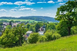 Eine malerische, ländliche Kleinstadt mit einem von Bäumen und Häusern umgebenen Kirchturm, eingebettet in sanfte grüne Hügel unter einem blauen Himmel mit vereinzelten Wolken. Wandern in der Oberlausitz.