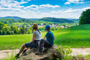 Zwei Kinder sitzen auf einem großen Stein und blicken auf eine malerische Landschaft mit grünen Feldern, sanften Hügeln und einer Kleinstadt - sie fangen den friedlichen Charme von Urlaub in der Oberlausitz unter einem strahlend blauen Himmel mit vereinzelten Wolken ein. Marktquartier 5 – Ferienapartments in Neusalza-Spremberg, Oberlausitz, Sachsen.