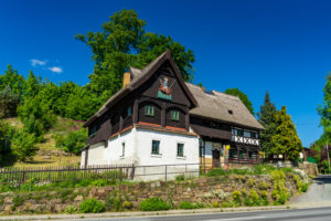 Das Reiterhaus, ein traditionelles Fachwerkhaus in der Oberlausitz, hat ein steiles Reetdach und hölzerne Akzente. Eingebettet hinter einer Steinmauer, inmitten von üppigem Grün und unter einem strahlend blauen Himmel, dient dieses historische Gebäude heute als charmantes Museum in Neusalza-Spremberg.
