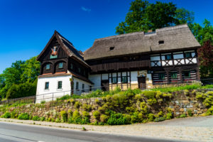 Das Reiterhaus, ein traditionelles Fachwerkhaus in der Oberlausitz, hat ein steiles Reetdach und hölzerne Akzente. Eingebettet hinter einer Steinmauer, inmitten von üppigem Grün und unter einem strahlend blauen Himmel, dient dieses historische Gebäude heute als charmantes Museum in Neusalza-Spremberg.