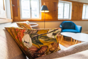 Nahaufnahme eines beigen Sofas mit einem blumengemusterten Kissen im gemütlichen Wohnzimmer im Ferienhaus Marktquartier 5 in der Oberlausitz. Im Hintergrund laden ein blauer Sessel, runde weiße Tische, eine Stehlampe und Fenster mit hölzernen Fensterläden dazu ein, den Urlaub zu genießen.