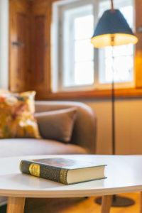 Ein geschlossenes Karl-May-Buch mit dekorativem Einband liegt auf einem weißen Tisch in einem gemütlichen Wohnzimmer, das Urlaubsgefühle hervorruft, mit einem Sofa, bunten Kissen, einer Stehlampe und einem sonnendurchfluteten Fenster im Hintergrund.