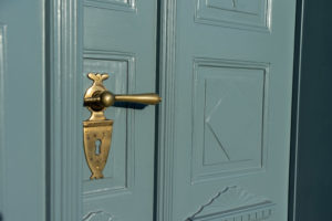 Nahaufnahme einer hellblauen Holztür mit dekorativen Paneelen und einem Messinggriff mit verziertem Schlüsselloch. Sonnenlicht wirft subtile Schatten auf die Türoberfläche. Zugang zum Ferienhaus Marktquartier 5.