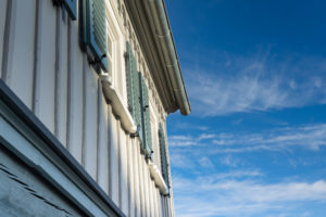 Nahaufnahme der Außenseite eines Hauses mit hellblauen Fensterläden und hellgrauer vertikaler Verkleidung, von unten vor einem strahlend blauen Himmel mit dünnen Wolken aufgenommen.