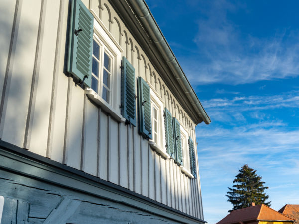Nahaufnahme eines weißen Holzhauses mit blaugrünen Fensterläden an den oberen Fenstern vor einem strahlend blauen Himmel mit feinen Wolken. Im Hintergrund sind ein weiteres Haus und eine hohe Fichte zu sehen.