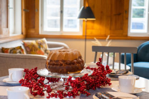 Ein Marmorkuchen auf einem Glasständer, umgeben von roten Beeren, steht auf einem gedeckten Tisch mit weißen Tassen und Untertassen in einer gemütlichen Ferienwohnung in der Oberlausitz, die mit Holzwänden und großen Fenstern ausgestattet ist.