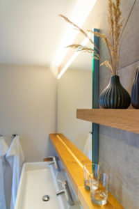Modernes Badezimmer mit weißem Waschbecken, Holzregal mit einer dunklen Vase mit getrockneten Gräsern, zwei transparenten Gläsern und einem Wandspiegel. Im Hintergrund hängen weiße Handtücher an Haken.