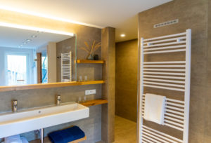 Modernes Badezimmer mit beige gefliesten Wänden, einem breiten weißen Waschbecken, einem großen Spiegel, Holzregalen mit Gläsern und dekorativen Zweigen, einem weißen Handtuchwärmer mit Handtuch und ordentlich gefalteten blauen Handtüchern auf einem Regal.
