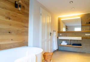 Modernes Badezimmer mit hölzerner Akzentwand, weißer Badewanne und doppeltem Waschtisch. Ideal für eine Ferienwohnung in der Oberlausitz. Ein großer beleuchteter Spiegel, Handtücher, einfaches Dekor und natürliches Licht durch ein Fenster sorgen für ein klares, minimalistisches Gefühl.