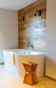 Ein modernes Badezimmer mit einer weißen freistehenden Badewanne vor einer holzgetäfelten Wand, zwei darüber angebrachten Wandleuchten, einem über der Wanne liegenden Handtuch und einem Holzhocker mit Seilakzenten im Vordergrund.