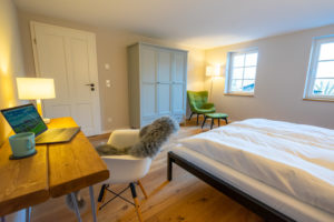 Ein gemütliches Schlafzimmer in einer Ferienwohnung mit Sauna, mit einem ordentlich gemachten Bett, einem Holzschreibtisch mit Laptop, Lampe und grüner Tasse, einem weißen Stuhl mit Pelzüberwurf, einem grünen Sessel, einem Kleiderschrank und zwei Fenstern, die den Raum mit natürlichem Licht füllen.